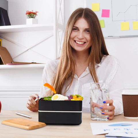 Woman smiling with Adult Bento Box With Compartments, 47oz Stackable Japanese Style Rectangle Lunch Container with utensils on desk.