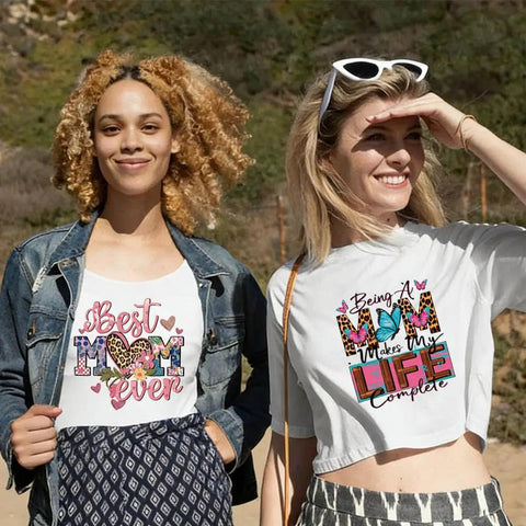 Two women wearing t-shirts with mom-themed designs including hearts and text outdoors.