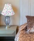 Floral lamp shade on clear glass table lamp next to bed with velvet brown bedding in cozy bedroom setting
