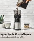 Barista Grade Conical Burr Electric Coffee Grinder With 12 Oz Capacity being filled with coffee beans next to a coffee cup