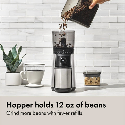 Barista Grade Conical Burr Electric Coffee Grinder With 12 Oz Capacity being filled with coffee beans next to a coffee cup
