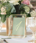Gold frame holding a table number card, surrounded by elegant glassware and floral centerpiece, used for events and celebrations.