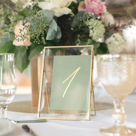 Gold frame holding a table number card, surrounded by elegant glassware and floral centerpiece, used for events and celebrations.