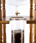Top Shelf Stainless Steel Cold Brew Dripper With Heavy Duty Premium Borosilicate Glass dripping coffee with wooden support.