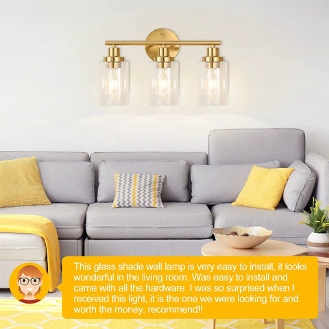 Brushed Brass 3 Light Vanity Wall Sconce With Clear Glass Shades, above gray and yellow sofa in living room.