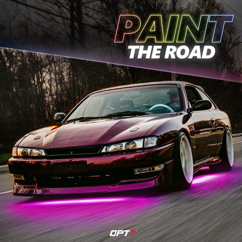 Car with pink Aura Pro Underglow LED Lights Kit on a road, showcasing neon underbody lighting and scenic background at dusk.
