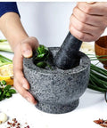 Hand using Mortar And Pestle Set Granite Spice Grinder 2 Cup Capacity with herbs, durable stone kitchen tool for home cooking.