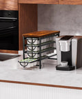 Universal Coffee Pod Holder With Two Multifunctional Storage Drawers For Tea Bags And K Cup Pods Nespresso Dolce Gusto CBTL Verismo Holds Up To 60 Pods In A Retro Brushed Bronze Finish displayed on a kitchen counter next to a coffee machine.