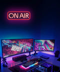 Gaming setup with dual monitors and bright 'ON AIR' neon sign wall decor illuminating room, contrasting colorful screen visuals.