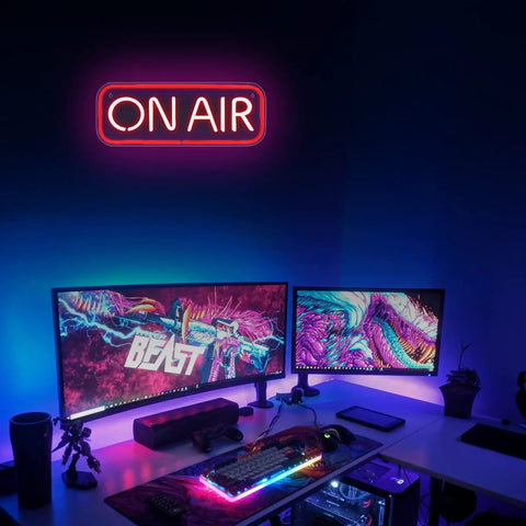Gaming setup with dual monitors and bright 'ON AIR' neon sign wall decor illuminating room, contrasting colorful screen visuals.