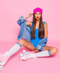Woman in pink skates with striped socks, wearing a pink beanie and blue outfit, sitting against a pink background.