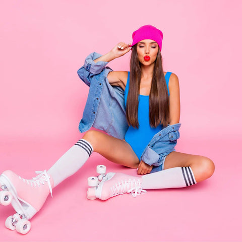 Woman in pink skates with striped socks, wearing a pink beanie and blue outfit, sitting against a pink background.