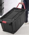 Person carrying Two Pack 160 Quart Black Stackable Rolling Storage Bins with Heavy Duty Red Latches and Smooth Glide Wheels.
