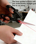 Side view of black laser-guided fabric scissors with ergonomic handles cutting white paper; red laser line visible.
