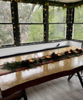 Decorated wooden dining table with holiday garland and candles, surrounded by windows and chairs, modern rustic setting.