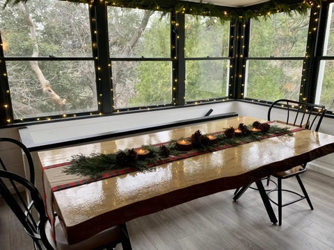 Decorated wooden dining table with holiday garland and candles, surrounded by windows and chairs, modern rustic setting.