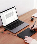 Compact Drawing Tablet With Slick Scroll Wheel And Ergonomic Stylus Delivering Sick Precision And Butter Smooth Pressure Response Perfect For Pc Artists Seeking Zero Lag Creativity And Hassle Free Setup In Sleek Black on a wooden desk next to a laptop.