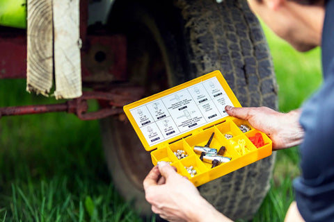 Yellow plastic grease fitting replacement kit case with SAE/metric zerks and caps, held near a wheel.
