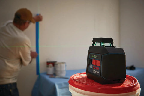 Green Beam Self Leveling 360 Degree Cross Line Laser With Batteries Included L Bracket Ceiling Clip And Hard Carrying Case For Pro Grade Horizontal And Vertical Layouts On The Job Site, placed on a construction bucket with worker in background