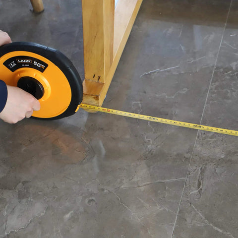 Person using 165 Feet Fiberglass Tape Measure With Dual Inch And Metric Scale under wooden table on tile floor