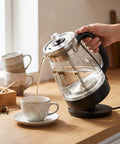 1 Liter Glass Electric Kettle With Keep Warm Function pouring hot tea into a white cup on a wooden table.
