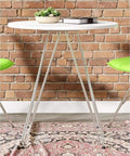 Round table with white top and slim metal legs on a patterned rug against a brick wall, green chairs partially visible.