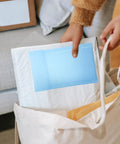 Person placing Milk Blue Bubble Mailer Bags 100 Pack Self Sealing Thickened 11x15 Plus 4 Inch Envelopes into a canvas bag