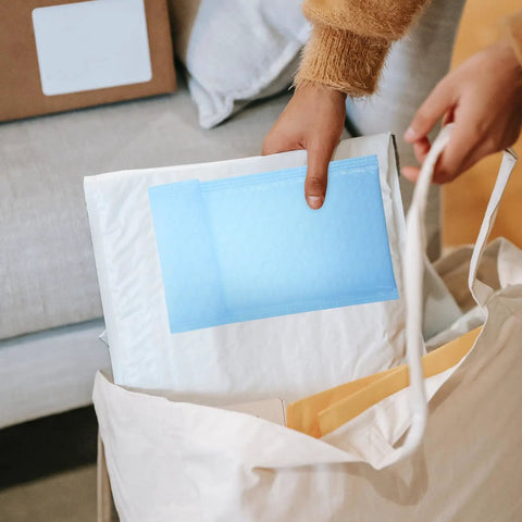Person placing Milk Blue Bubble Mailer Bags 100 Pack Self Sealing Thickened 11x15 Plus 4 Inch Envelopes into a canvas bag