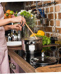 Person cooking with a stainless steel pot on a stovetop, surrounded by fresh vegetables and kitchen utensils.