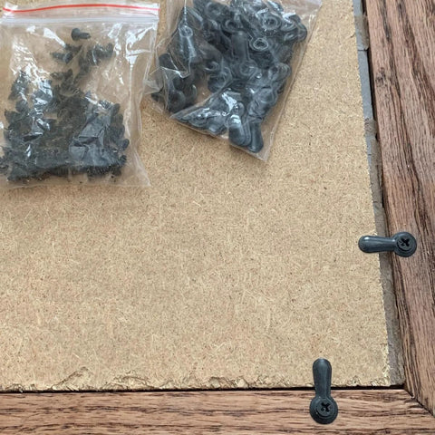 Picture frame hardware kit with 100 black backing clips and turn-buttons with screws, shown on wooden-framed corkboard.