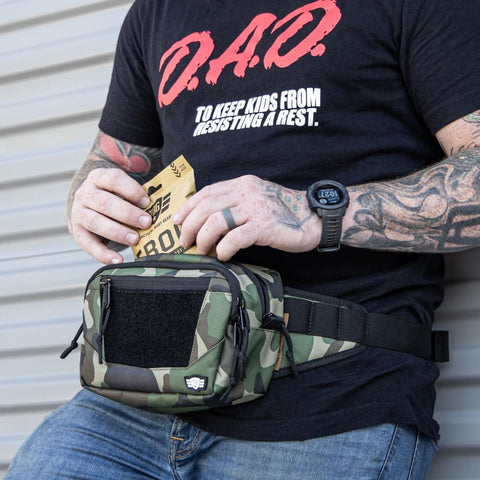 Person holding an emergency diaper kit with a camo waist pack, wearing a D.A.D. shirt, highlighting on-the-go parent convenience.
