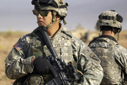 Two soldiers in camouflage uniforms with tactical gear and helmets, one holding a rifle in a desert setting.