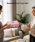 Two women exchanging gift boxes in a living room with a pink box labeled 'Sweet Gift for Your Girl' on the table.
