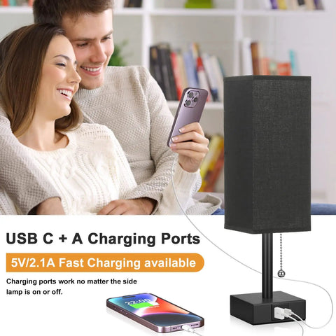 Black Bedside Table Lamp With USB C And USB A Ports, 3 Brightness Levels, shown on a table next to a couple with a smartphone.