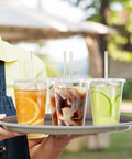Person holding tray with 100 Pack 16 Oz Clear Plastic Cups with Lids and Straws, various cold drinks visible