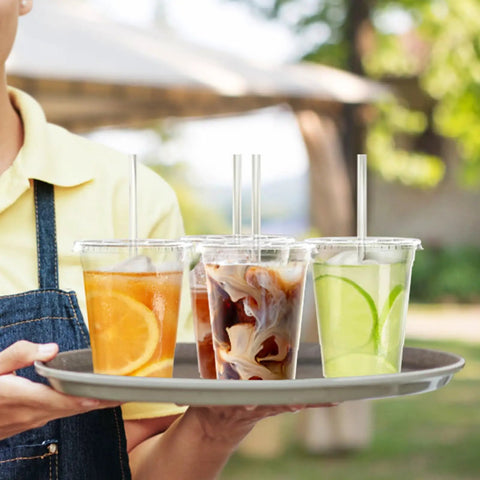 Person holding tray with 100 Pack 16 Oz Clear Plastic Cups with Lids and Straws, various cold drinks visible