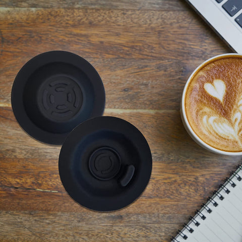 Two black 54mm espresso backflush cleaning discs for Breville machines on a wooden table, beside a latte and notebook
