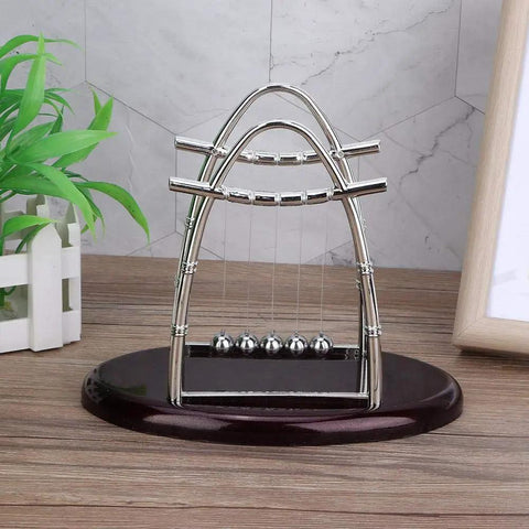 Pendulum Ball Desk Decor Set That Demonstrates Newton's Laws With Swinging Balls on wooden base with greenery in background