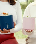Two women holding food storage jars, a blue and a pink version, showcasing the Slick Rugged Badass 25 Oz Double Wall Vacuum Insulated Food Storage Jar With Wide Mouth Leak Proof Lid Keeps Your Grub Scalding Hot Ten Hours Or Ice Cold Sixteen Hours Matte Black.