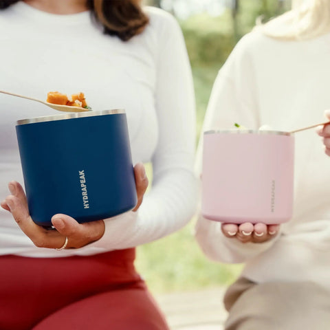 Two women holding food storage jars, a blue and a pink version, showcasing the Slick Rugged Badass 25 Oz Double Wall Vacuum Insulated Food Storage Jar With Wide Mouth Leak Proof Lid Keeps Your Grub Scalding Hot Ten Hours Or Ice Cold Sixteen Hours Matte Black.