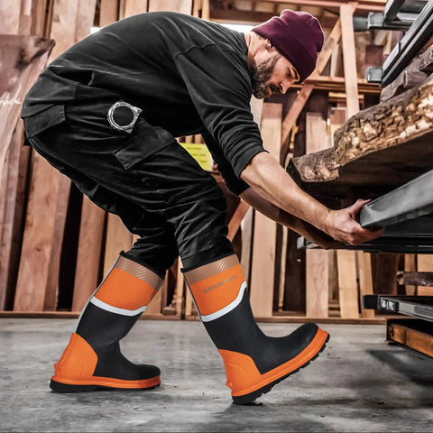 Person wearing Steel Toe Rubber Boots For Men With Puncture Proof Steel Shank Waterproof Insulated 6mm Neoprene Work Boots With Reflective Strips For Farming Manufacturing And Construction Black Orange Size 8, shown in a workshop setting.
