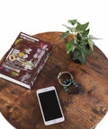 Round Two Tier Side Table With Open Storage Shelf, Anti Scratch Protective Pads, Kid Safe Curved Edges, Straight Up Sturdy Metal End Table For Cozy Living Rooms And Bedrooms, featuring books, phone, and plant on rustic brown surface.