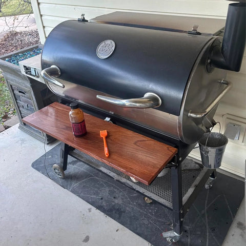 Pellet grill with condiment bottle and brush on a wooden side shelf, outdoor setting.