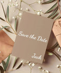 Save the date card with name Jack on beige paper, surrounded by envelopes and flowers.