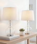 25 Inch Clear Chrome And Brass Glass Table Lamps Set Of 2 With White Linen Drum Shades on a Wooden Table