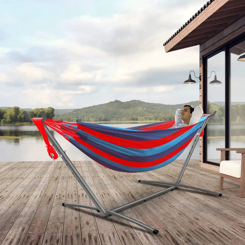 Man relaxing in Easy Setup Brazilian Style Hammock With Tree Straps For Hanging, Blue And Red Stripe, on patio by lake.