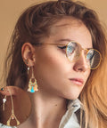 Woman wearing glasses with a gold-colored earring made from 40Pcs Stainless Steel Earring Hooks Hypoallergenic V Shaped Ear Wires And 40Pcs Clear Bullet Earring Backs For DIY Dangle Earrings Jewelry Making Two Color Set Replacement Kit Must Have.