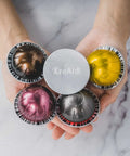 Hands holding reusable aluminum foil lids for Nespresso VertuoLine pods with assorted coffee capsules, showcasing design and color.