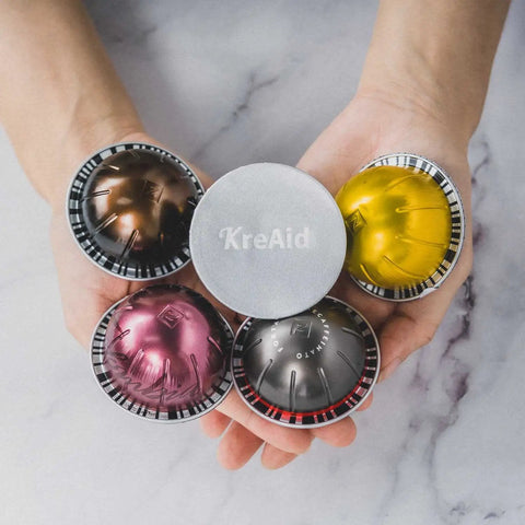 Hands holding reusable aluminum foil lids for Nespresso VertuoLine pods with assorted coffee capsules, showcasing design and color.