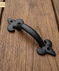 Old School Medieval Rustic Cast Iron Door Pull With Fleur De Lis Handle And Compact Grip, 7 1/8 Inch on wood surface